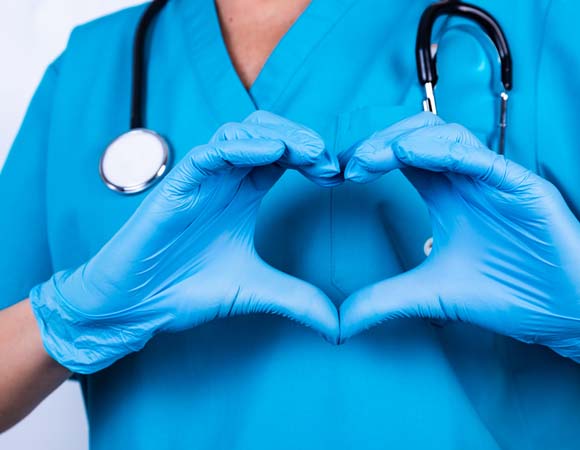 person in latex gloves, scrubs and a stethoscope holds up a heart sign with their hands over their heart